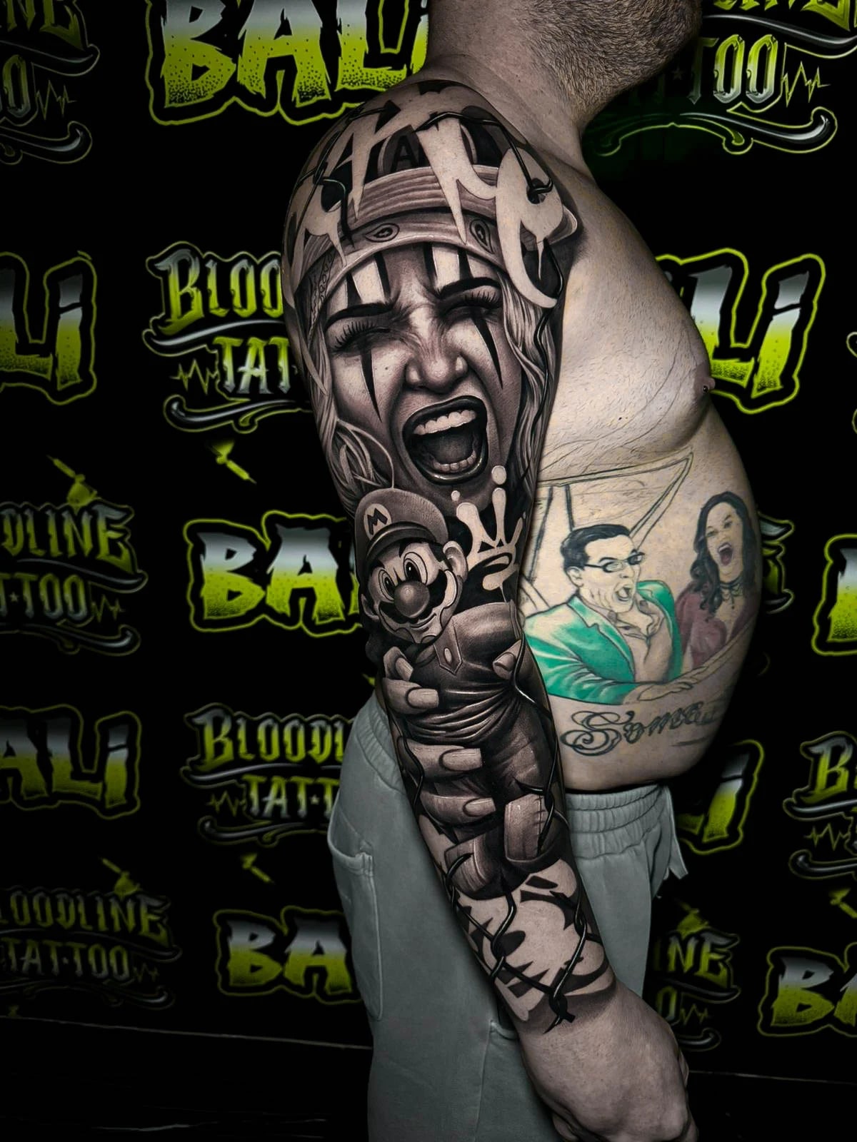 Detailed black and grey Chicano upper arm sleeve tattoo featuring a screaming clown girl, Mario figure, and bold graffiti-style script at Bloodline Tattoo Bali.