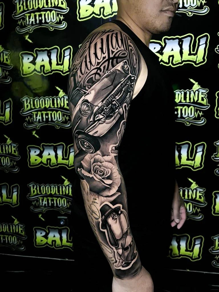 Detailed black and grey Chicano full arm sleeve tattoo with a lowrider car, rose, script lettering, and a suited penguin character at Bloodline Tattoo Bali.