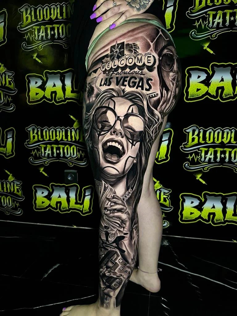Detailed black and grey Chicano thigh tattoo with the Las Vegas sign, laughing woman in round glasses, skull, and gambling motifs at Bloodline Tattoo Bali.