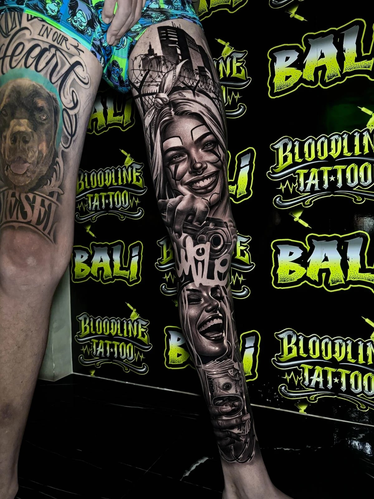 Detailed black and grey Chicano leg sleeve tattoo with smiling clown women, a handgun, city skyline, and stacked portrait faces at Bloodline Tattoo Bali.