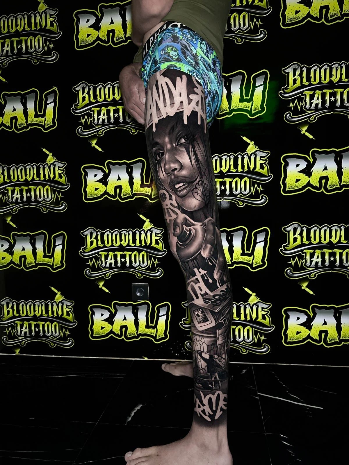 Detailed black and grey Chicano leg sleeve tattoo featuring a female portrait, spray can, block lettering, and urban street imagery at Bloodline Tattoo Bali.
