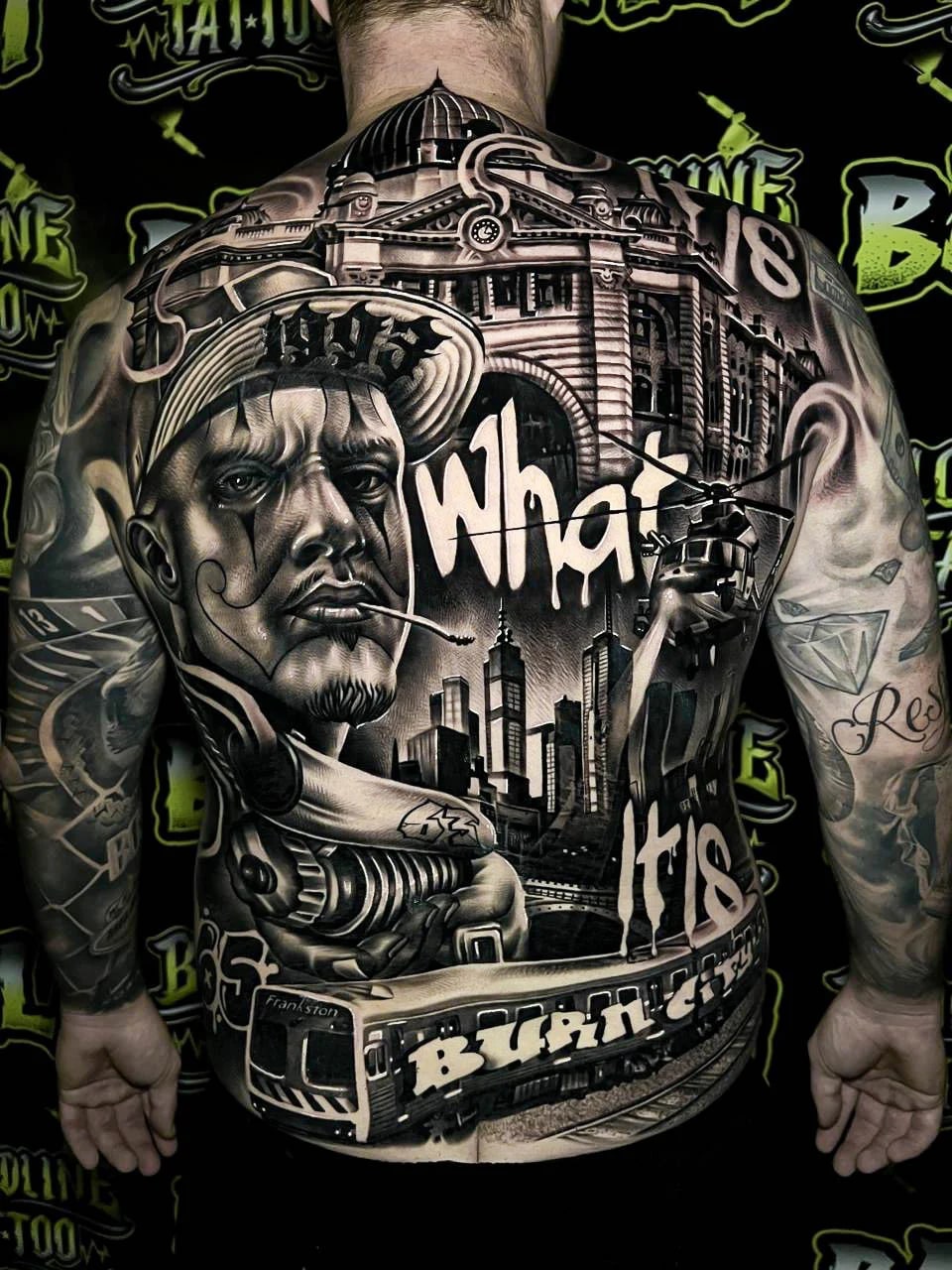 Detailed black and grey Chicano full back tattoo with a gangster portrait, city skyline, helicopter, tram, and large script lettering at Bloodline Tattoo Bali.