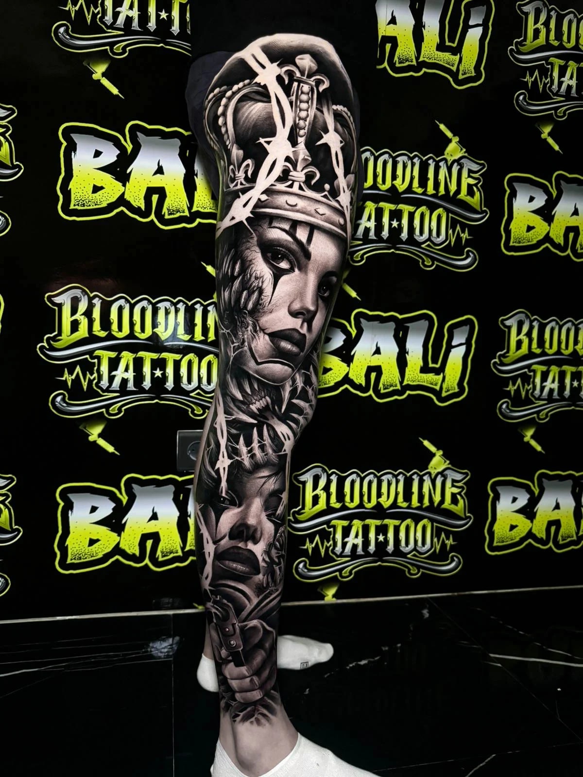 Detailed black and grey Chicano leg sleeve tattoo featuring a crowned woman portrait, barbed wire elements, layered faces, and a pistol at Bloodline Tattoo Bali.