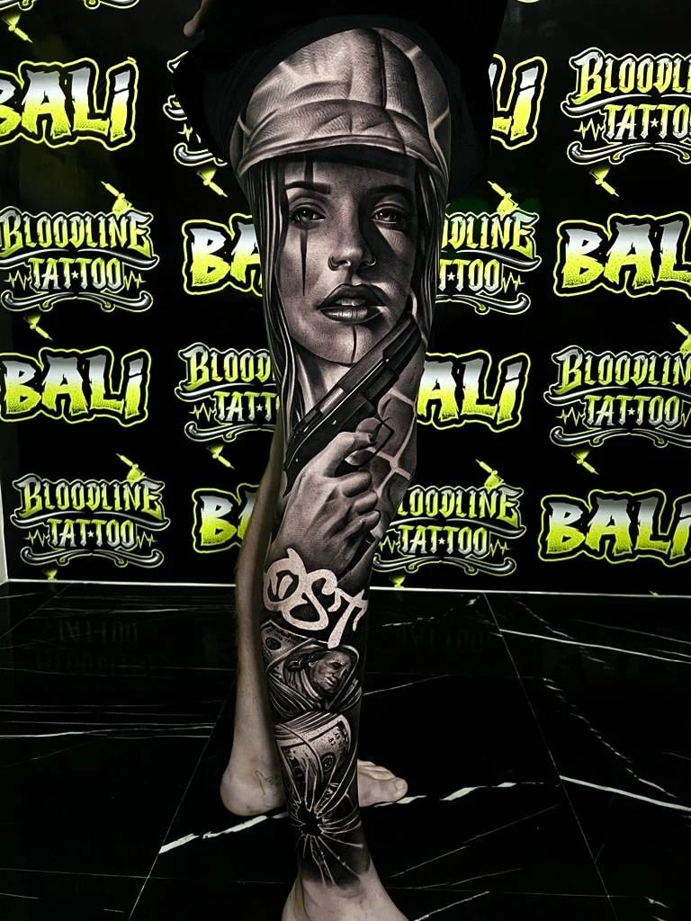 Detailed black and grey Chicano leg sleeve tattoo of a woman in a cap holding a pistol with cash and a nun portrait below at Bloodline Tattoo Bali.