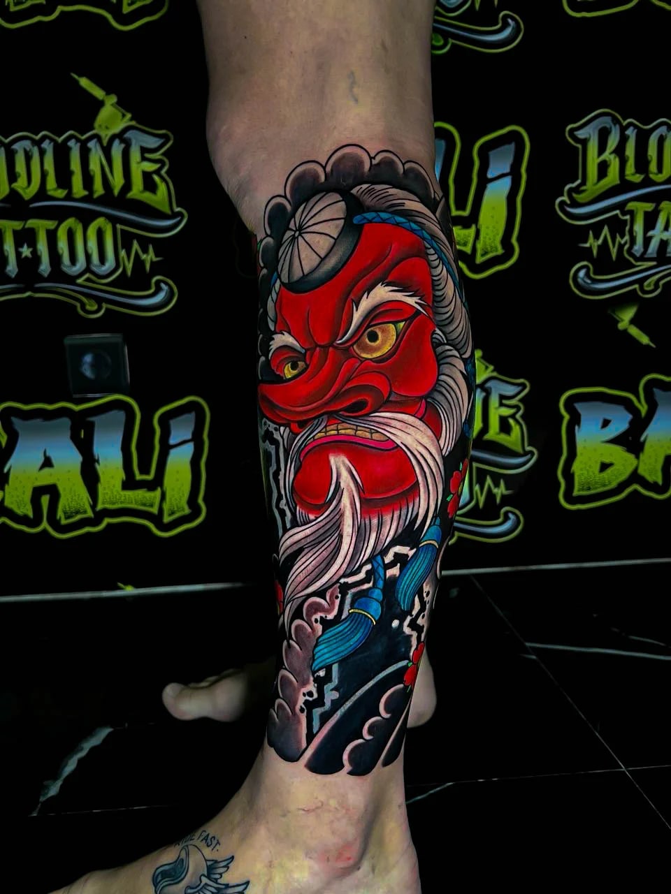 Colour Japanese calf tattoo of a fierce red daruma face with blue scarf details and black wave shading at Bloodline Tattoo Bali.