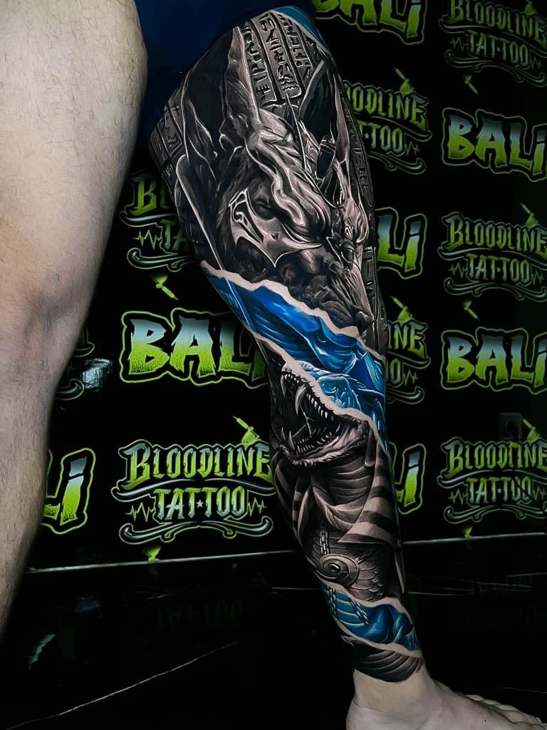 Colour fantasy leg sleeve tattoo with a stone dragon, blue torn-water effect, and a snarling shark breaking through below at Bloodline Tattoo Bali.