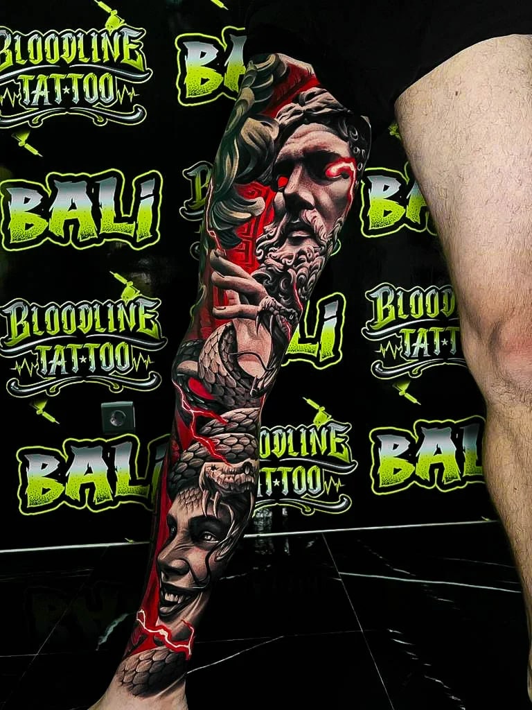 Colour leg sleeve tattoo featuring a red-eyed classical statue, coiling snake scales, and a sinister grinning face at Bloodline Tattoo Bali.