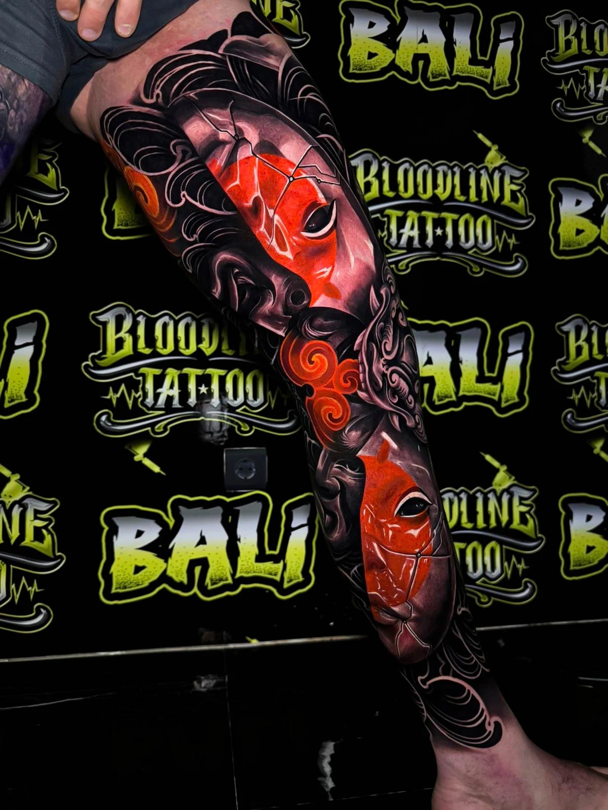 Colour thigh tattoo with cracked red theatrical masks, flowing black hair forms, and glowing orange cloud accents at Bloodline Tattoo Bali.