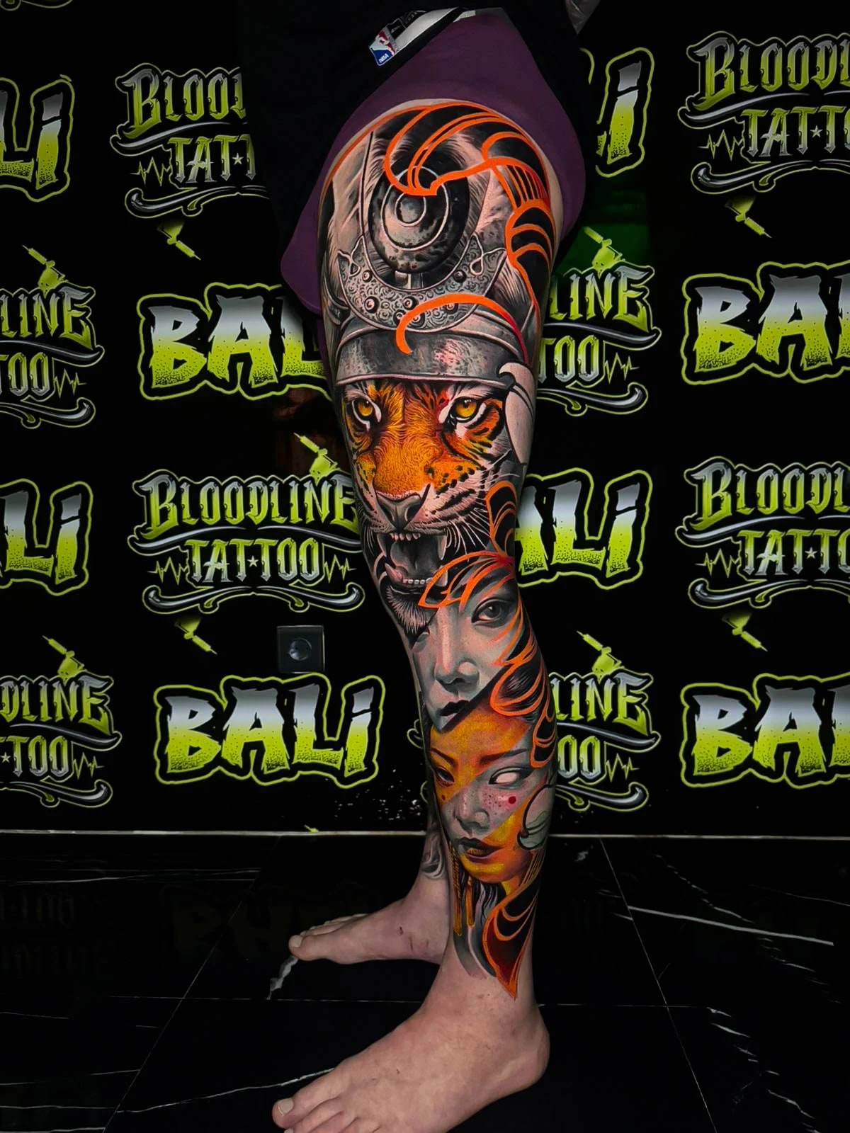 Colour leg sleeve tattoo with a roaring tiger in a samurai helmet, soft female portraits, and orange ornamental swirls at Bloodline Tattoo Bali.