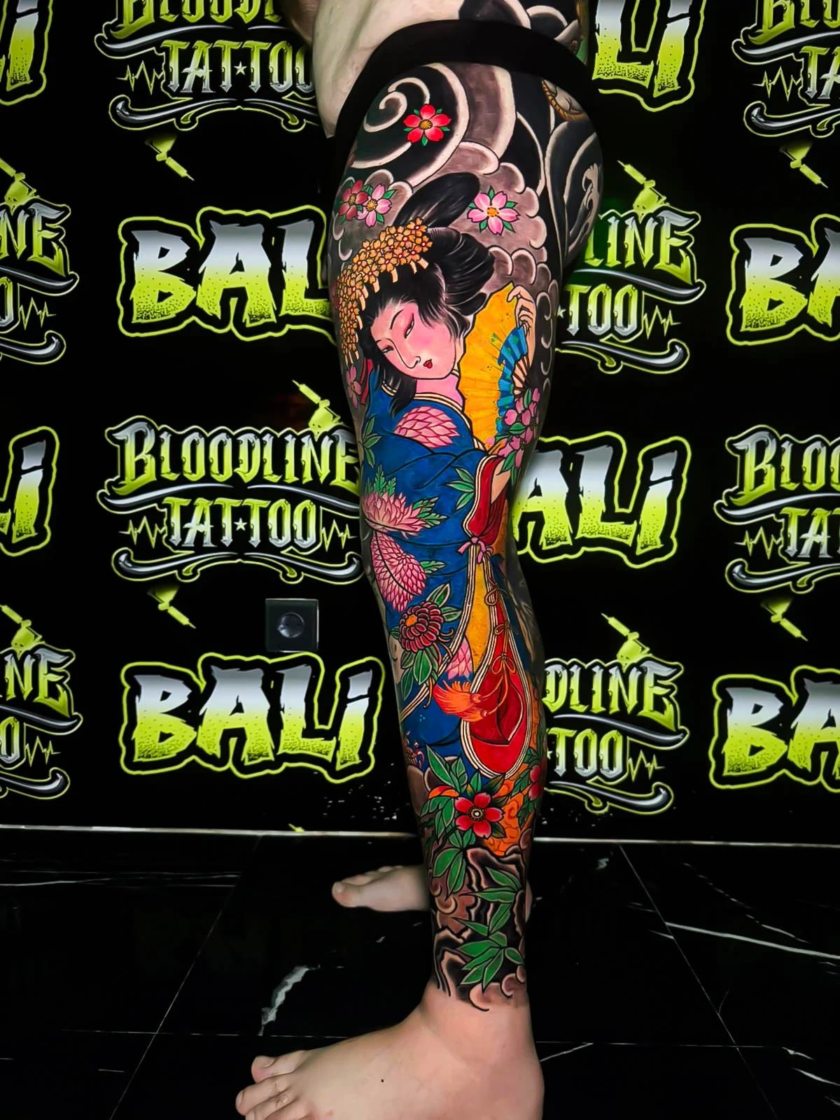 Colour Japanese leg sleeve tattoo of a geisha in a blue kimono surrounded by blossoms, leaves, and traditional black cloud shading at Bloodline Tattoo Bali.