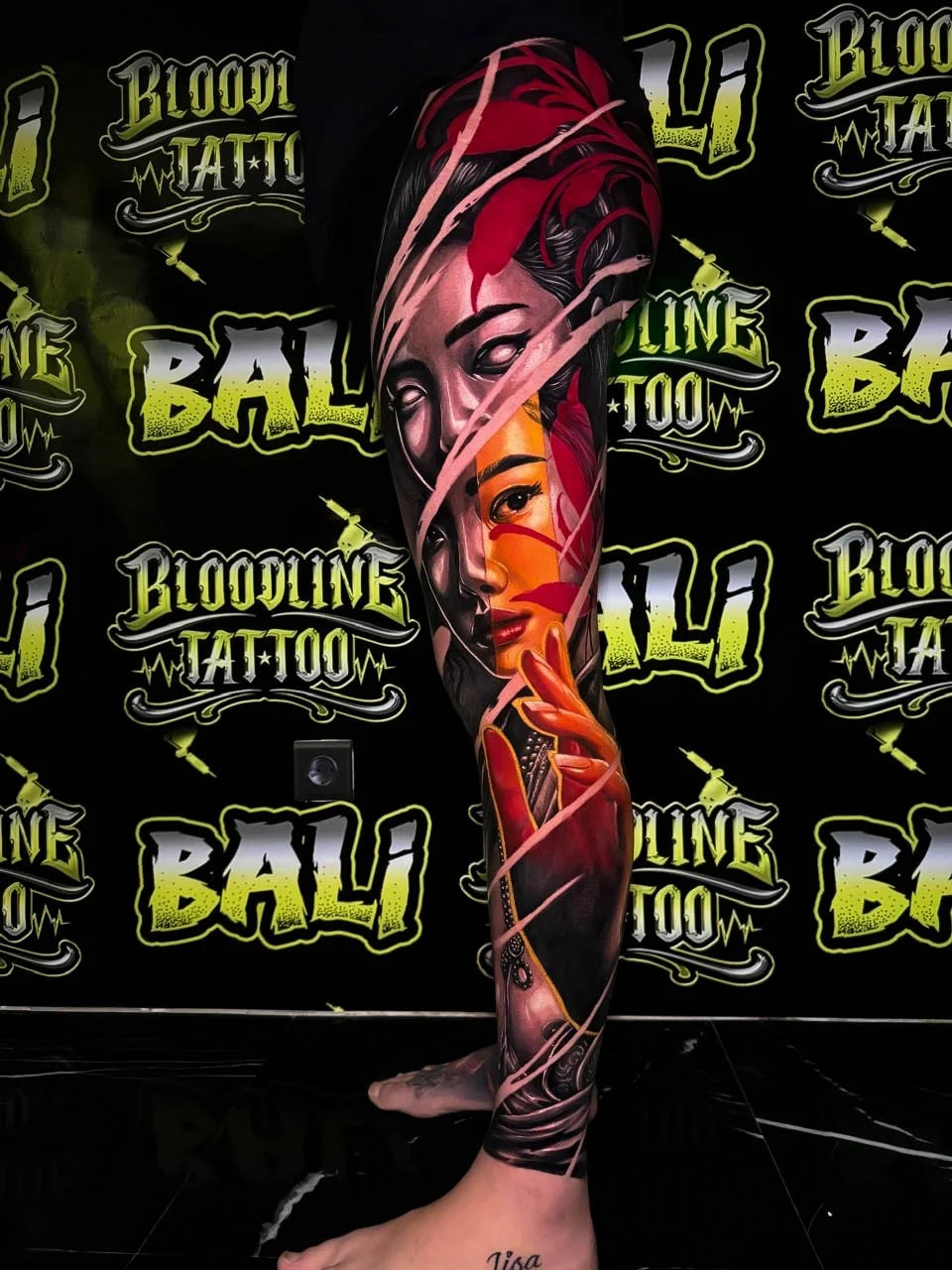Colour surreal leg tattoo with layered female faces, red ribbon slashes, and orange highlights flowing through the composition at Bloodline Tattoo Bali.