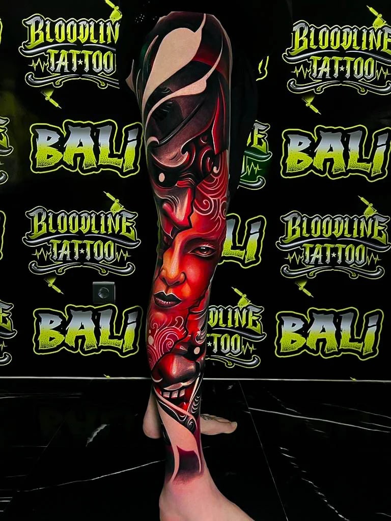 Colour abstract leg sleeve tattoo with stacked crimson female faces, black helmet shapes, and flowing ornamental red linework at Bloodline Tattoo Bali.