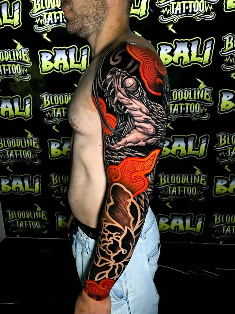 Colour upper arm sleeve tattoo of a roaring cobra head with bold blackwork, orange clouds, and floral negative-space shapes at Bloodline Tattoo Bali.