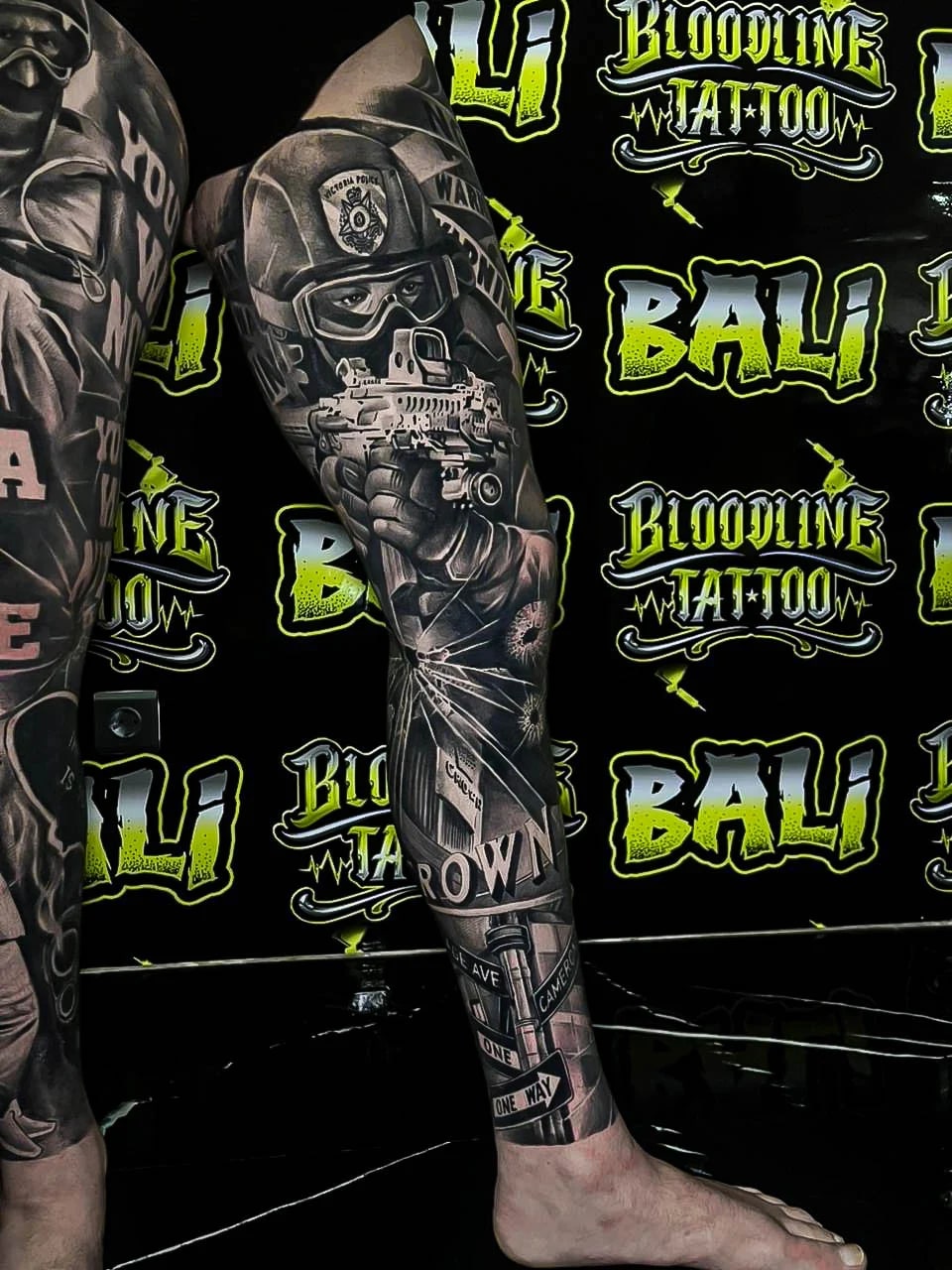 Healed realism leg sleeve with an armed officer, street signs, and shattered glass, keeping sharp healed detail and solid black-and-grey contrast at Bloodline Tattoo Bali.