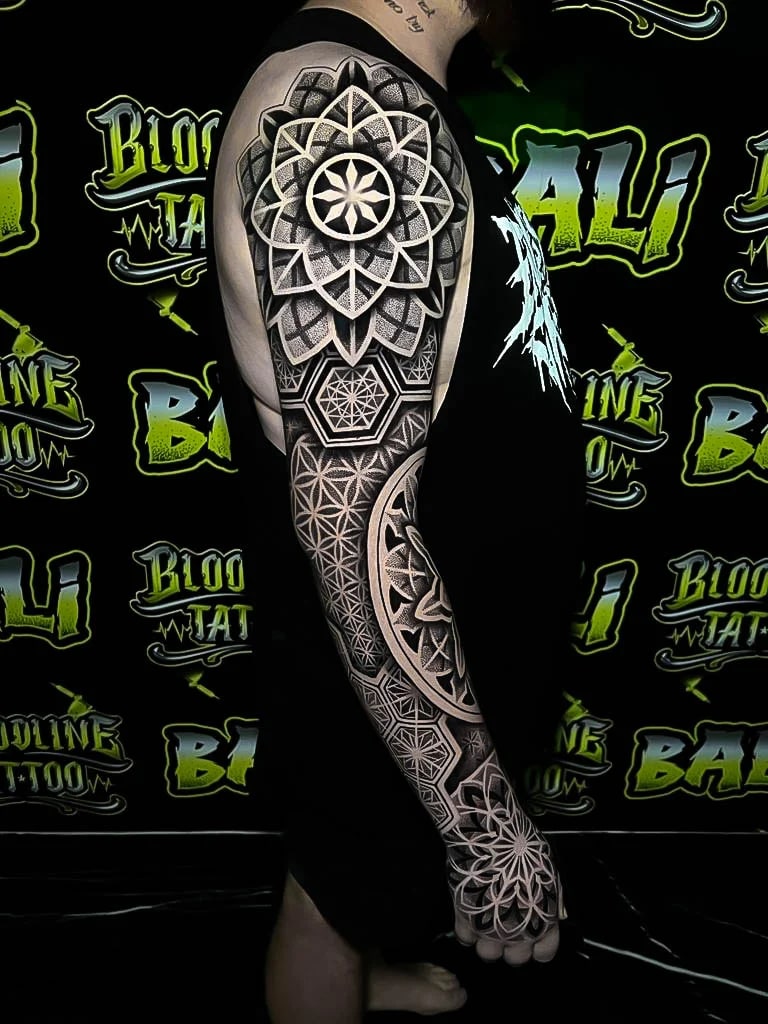 Full sleeve with a shoulder mandala, Flower of Life panels, geometric hand rosette, and saturated blackout contrast through the arm at Bloodline Tattoo Bali.