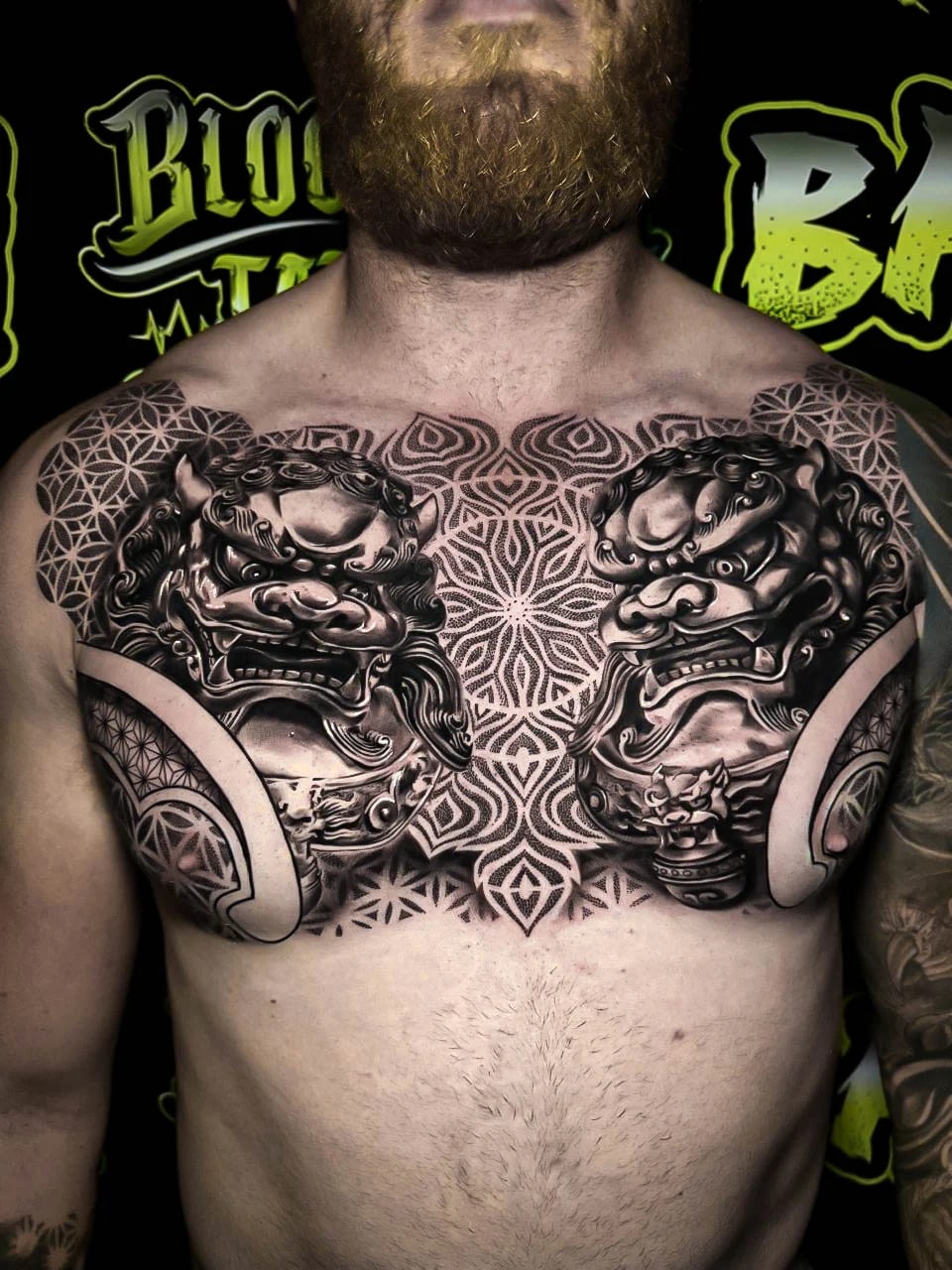 Chest piece with mirrored guardian lions, a central mandala bloom, and Flower of Life shoulders rendered in smooth black-and-grey at Bloodline Tattoo Bali.