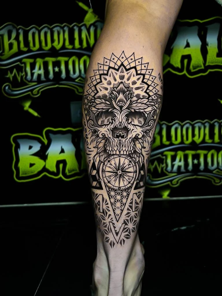 Calf piece with a skull, lotus bloom, compass dial, and geometric dotwork panels stacked into a sharp black-and-grey composition at Bloodline Tattoo Bali.