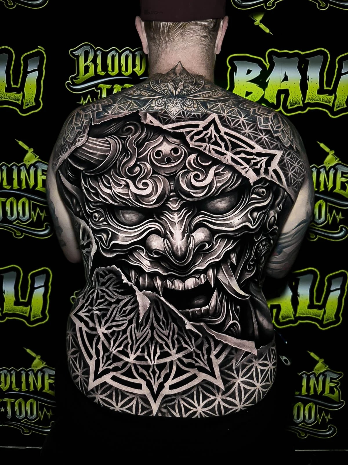 Full-back black-and-grey Oni reveal with torn geometric framing, Flower of Life shoulders, and bold ornamental mandala work across the lower back at Bloodline Tattoo Bali.