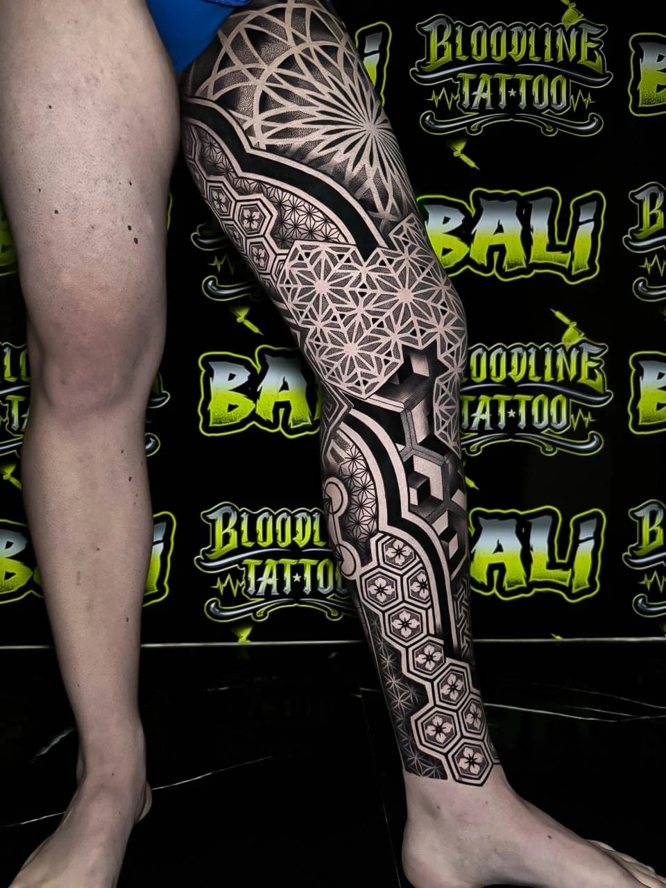 Side leg sacred geometry with a large rosette fan, clustered Flower of Life cells, and cube-grid blackwork cut with clean negative space at Bloodline Tattoo Bali.