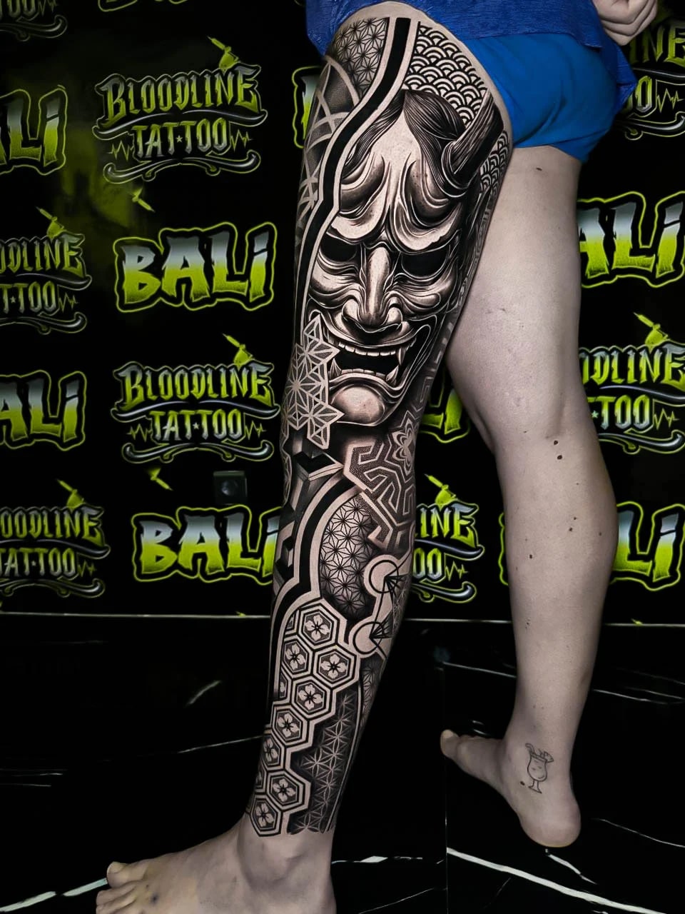 Leg sleeve with a Hannya face, honeycomb tiles, wave fill, and Flower of Life geometry layered into smooth black-and-grey shading at Bloodline Tattoo Bali.