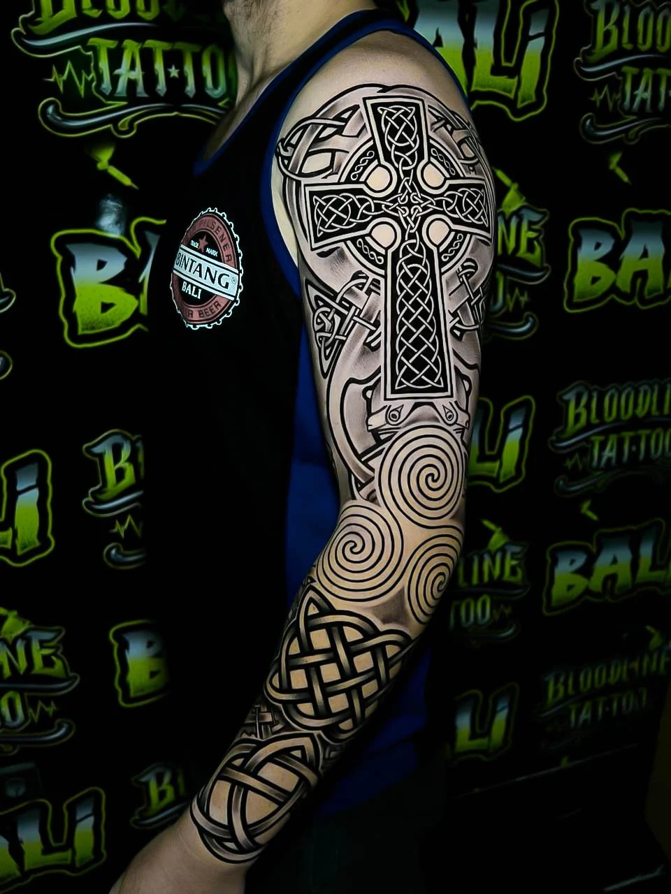 Blackwork sleeve with a Celtic cross, spiral triskeles, and knotwork bands flowing from shoulder to forearm in clean high-contrast linework at Bloodline Tattoo Bali.