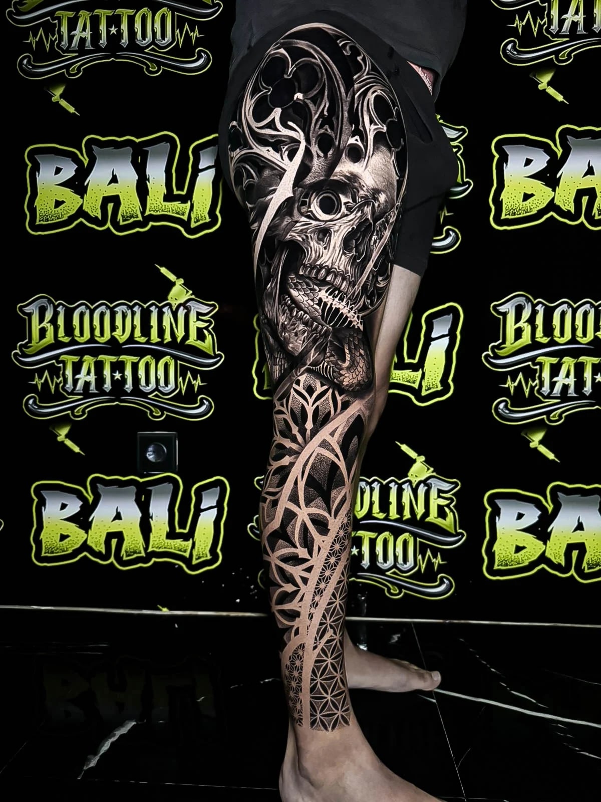 Leg sleeve with a gothic skull, cathedral-style tracery, and Flower of Life geometry descending into the shin in smoky black-and-grey at Bloodline Tattoo Bali.