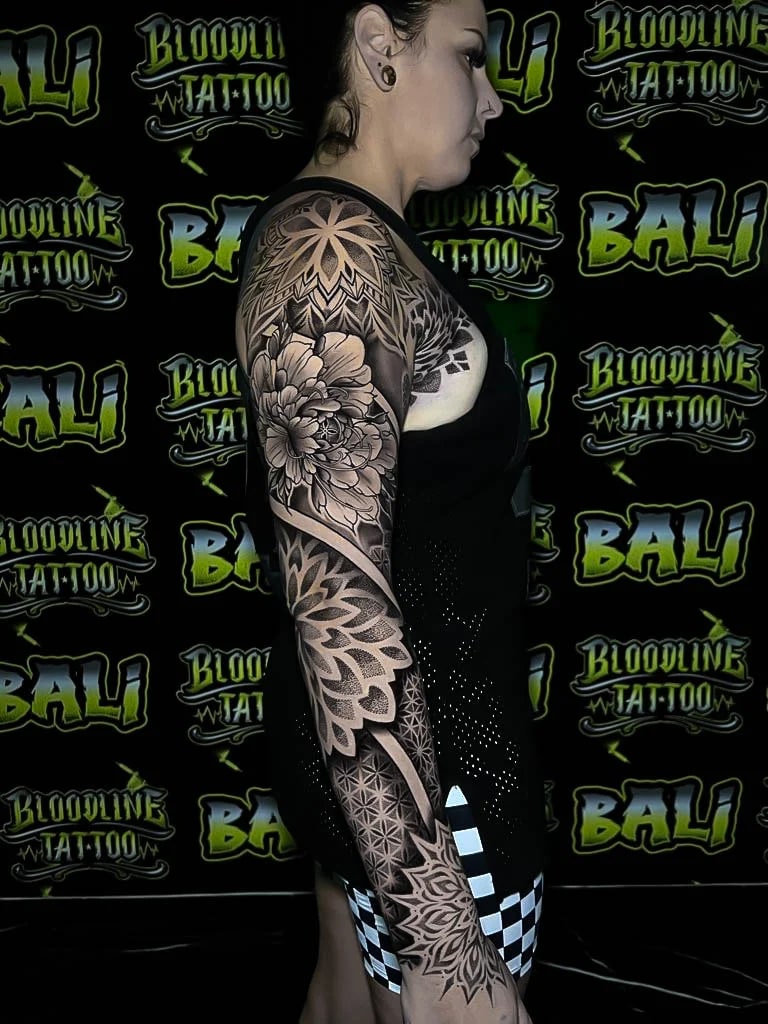 Black-and-grey sleeve with a shoulder starburst mandala, peony bloom, and geometric dotwork running into ornamental wristwork at Bloodline Tattoo Bali.
