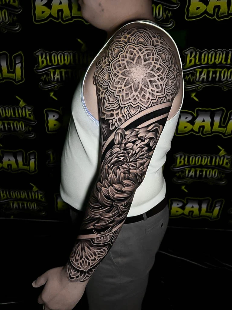 Upper-arm mandala cap with layered petals, a black-and-grey chrysanthemum, and ornamental wrist geometry finished in smooth dot shading at Bloodline Tattoo Bali.