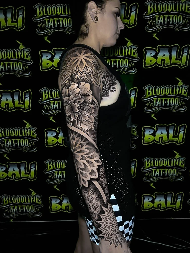 Black-and-grey sleeve with a shoulder starburst mandala, peony bloom, and geometric dotwork running into ornamental wristwork at Bloodline Tattoo Bali.