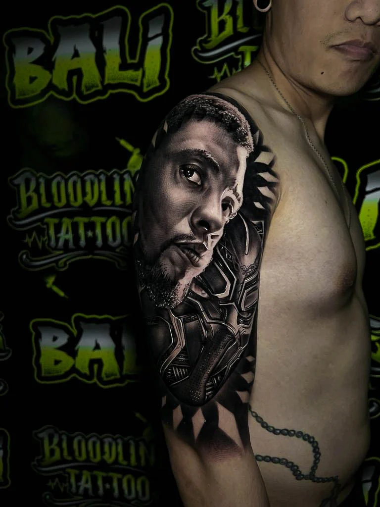 Black and grey portrait realism shoulder tattoo of Chadwick Boseman as Black Panther with a detailed superhero mask background at Bloodline Tattoo Bali.