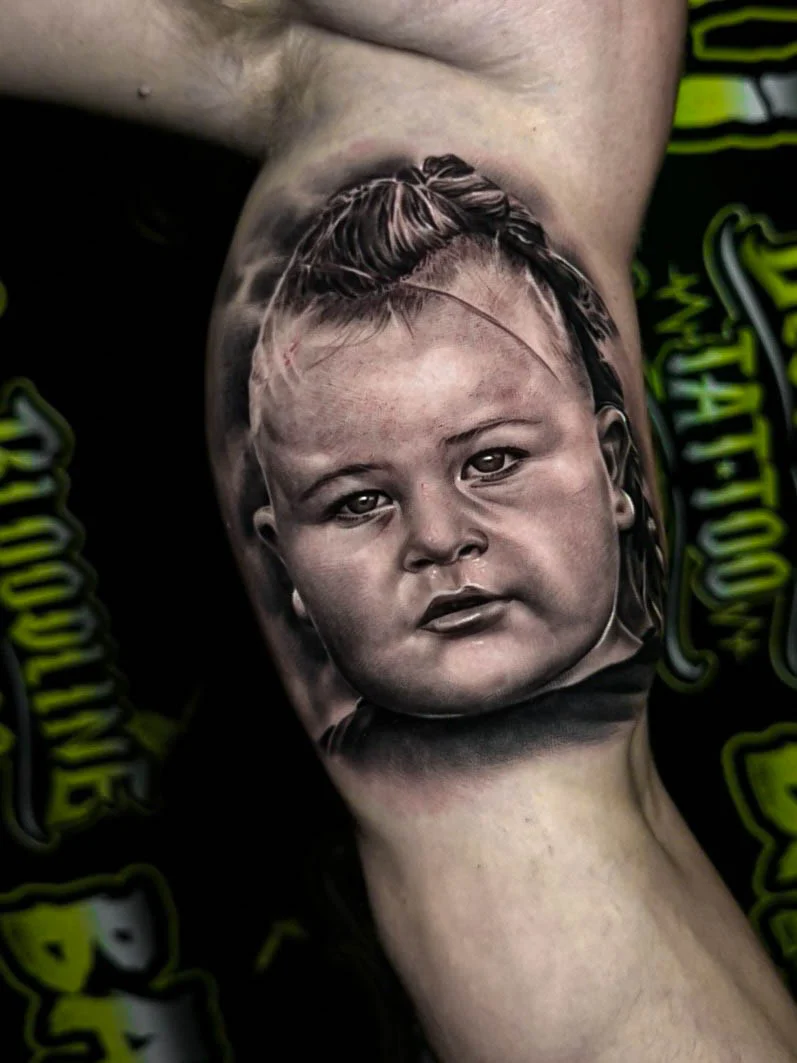 Black and grey portrait realism tattoo of a baby face with soft skin shading and detailed eyes at Bloodline Tattoo Bali.