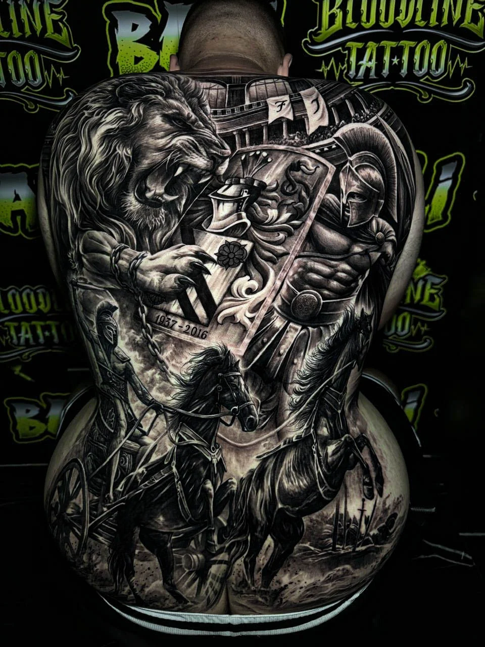 A black and grey realism full back piece with a roaring lion, armored warrior figures, and battle scene textures in dramatic contrast, at Bloodline Tattoo Bali.