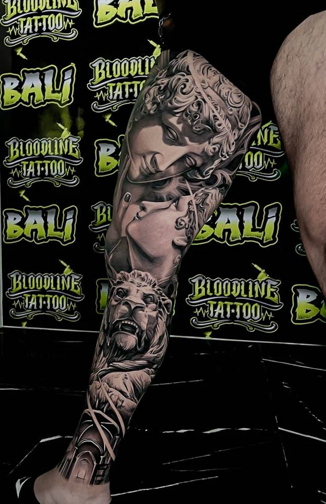 A black and grey realism upper arm sleeve with Scarface, a femme fatale, a handgun, and cash layered in gritty gangster shading, at Bloodline Tattoo Bali.