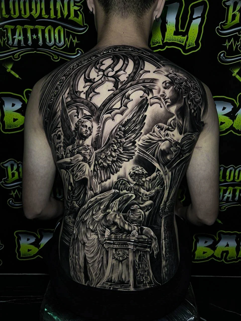 A black and grey realism full back piece with a winged angel, moonlit clouds, and cathedral-inspired sacred imagery in cinematic shading, at Bloodline Tattoo Bali.
