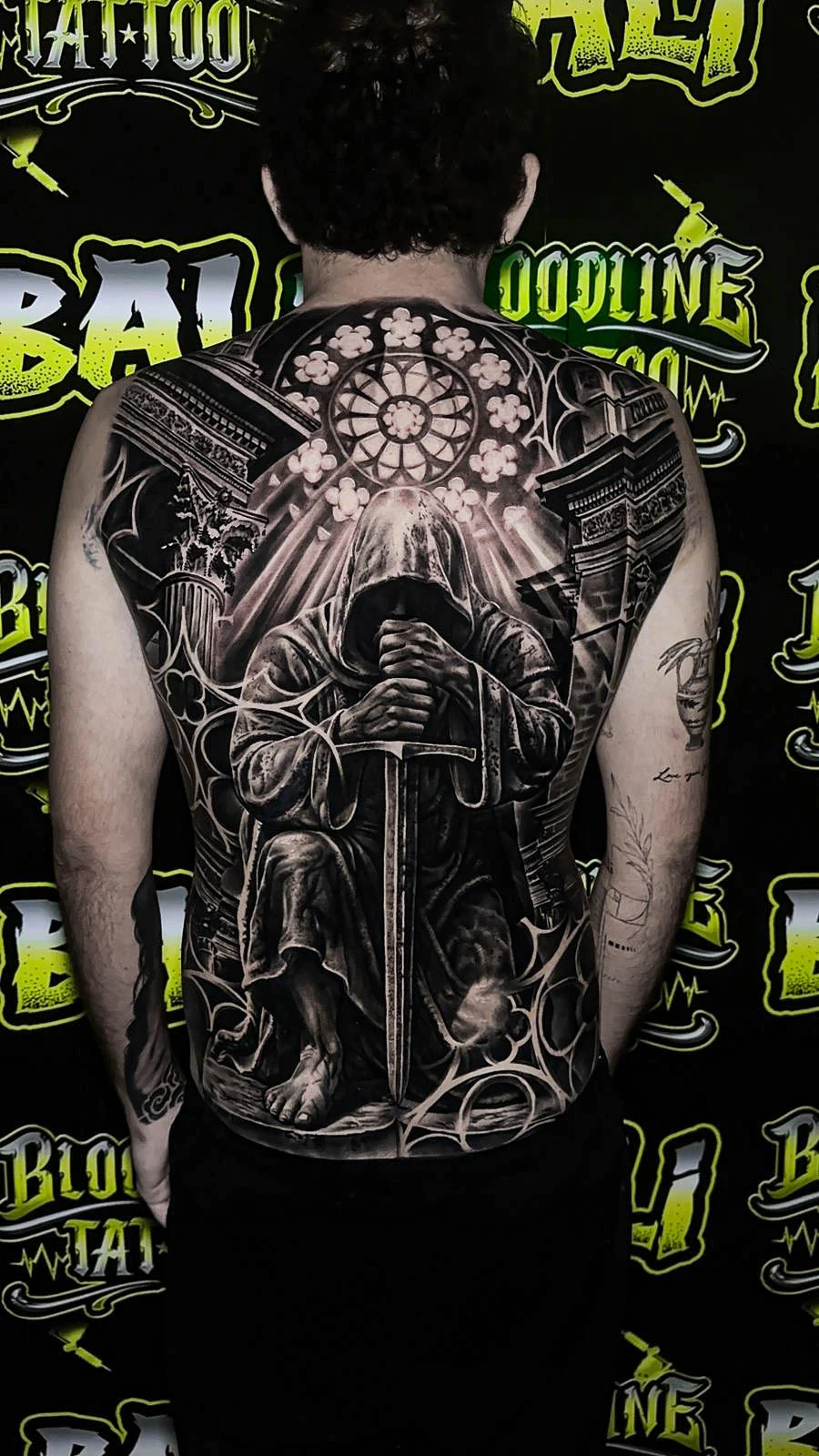 A black and grey realism full back piece of a grim reaper gripping a sword beneath stained-glass cathedral geometry, at Bloodline Tattoo Bali.