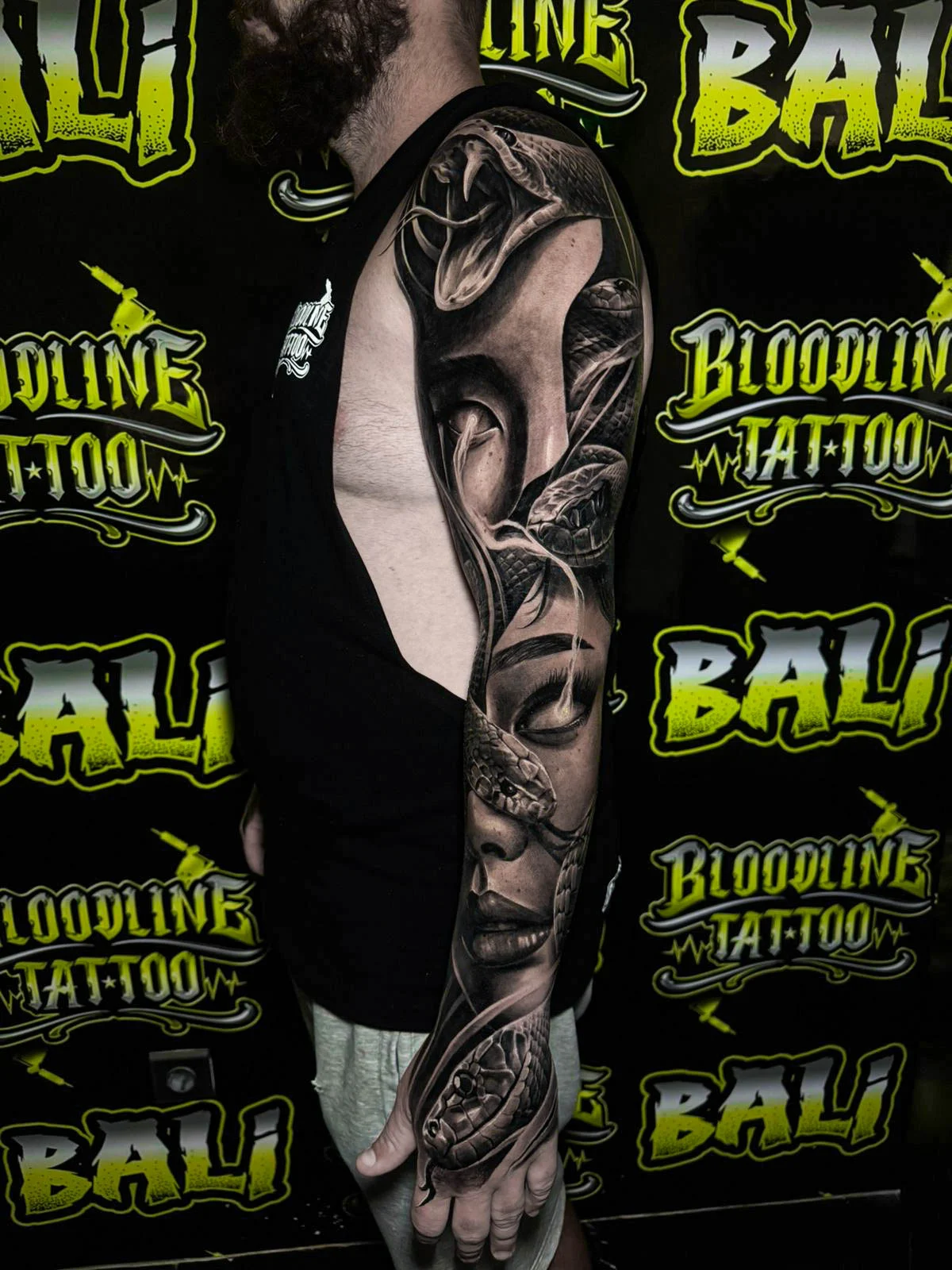 A black and grey realism arm sleeve with a split female face, skeletal detail, and smooth black and grey transitions through the upper arm, at Bloodline Tattoo Bali.