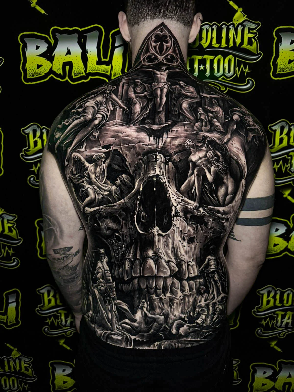 A black and grey realism full back piece with a giant skull, hooded figures, and battlefield-like textures in dramatic contrast, at Bloodline Tattoo Bali.