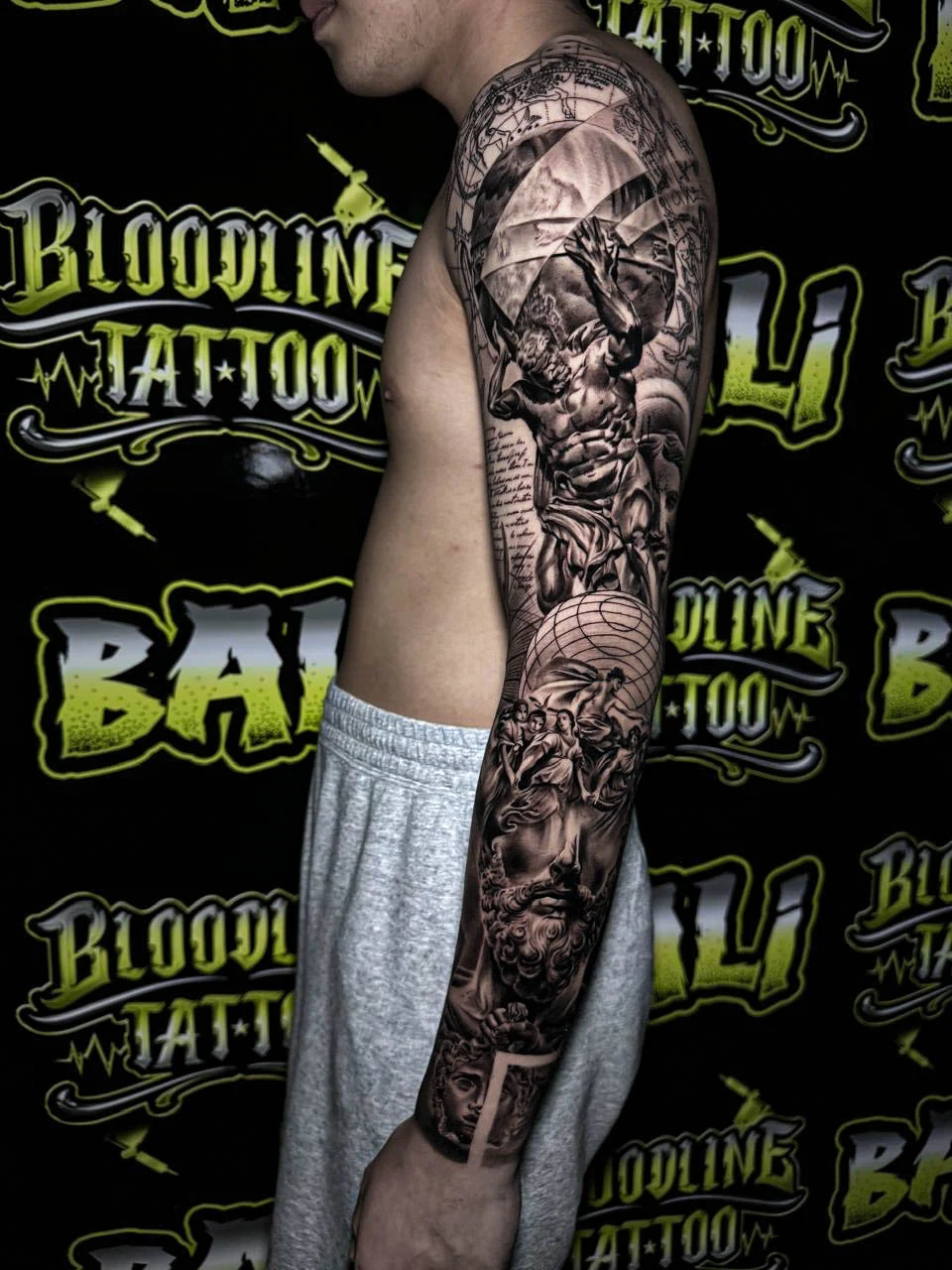 A black and grey realism arm sleeve with layered warrior imagery, textured faces, and rich black and grey detail wrapping the upper arm, at Bloodline Tattoo Bali.
