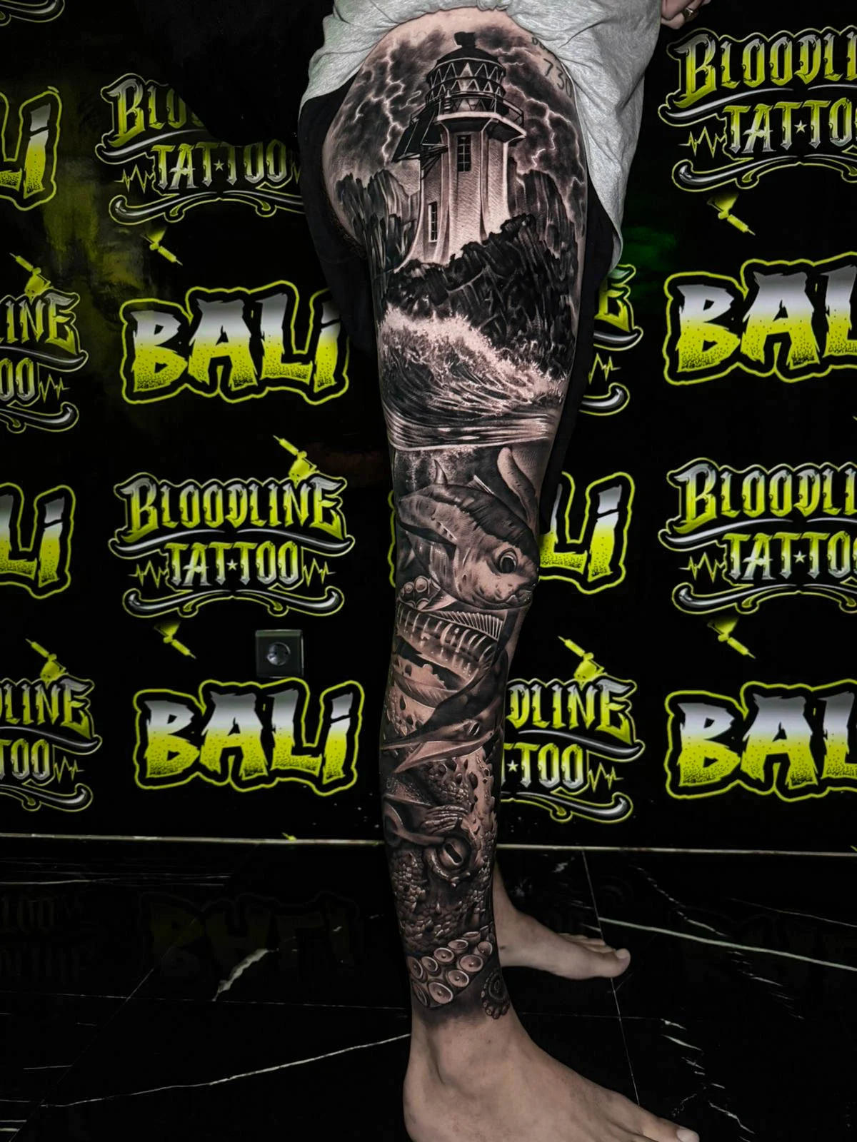 A black and grey realism lower leg sleeve with layered landscape imagery, dark textures, and soft smoke shading down the calf, at Bloodline Tattoo Bali.